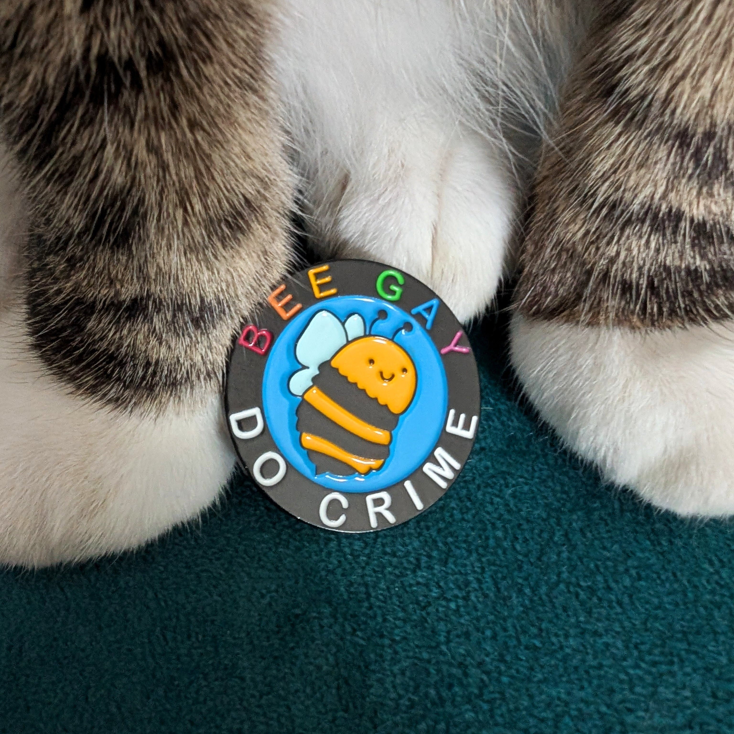 A close-up of a round enamel pin featuring a cartoon bee and the text 'BEE GAY DO CRIME' on a dark gray background, positioned between a cat's paws on a teal surface.