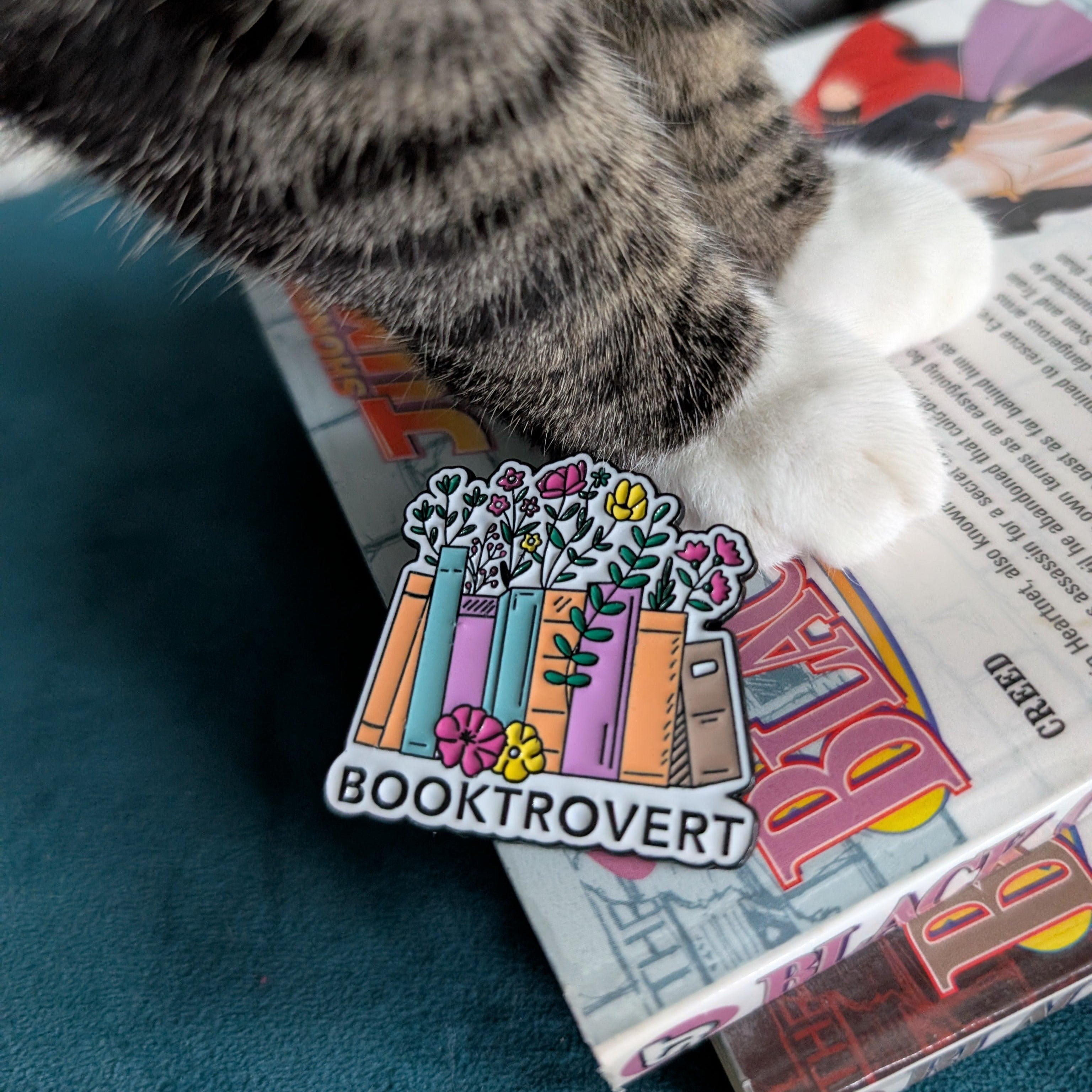 Cat paw with a 'Booktrovert' enamel pin on a book
