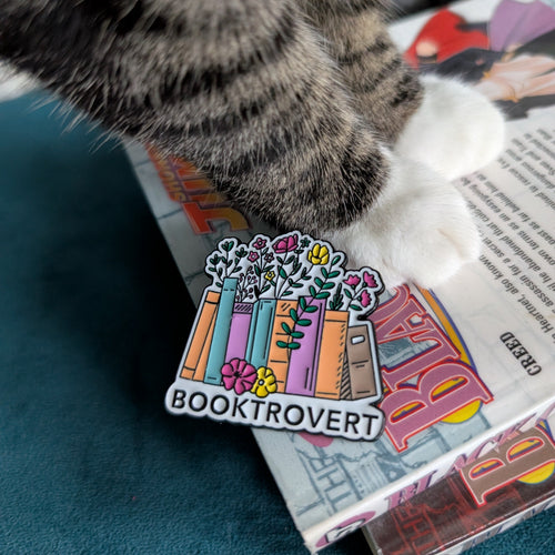 Cat paw with a 'Booktrovert' enamel pin on a book