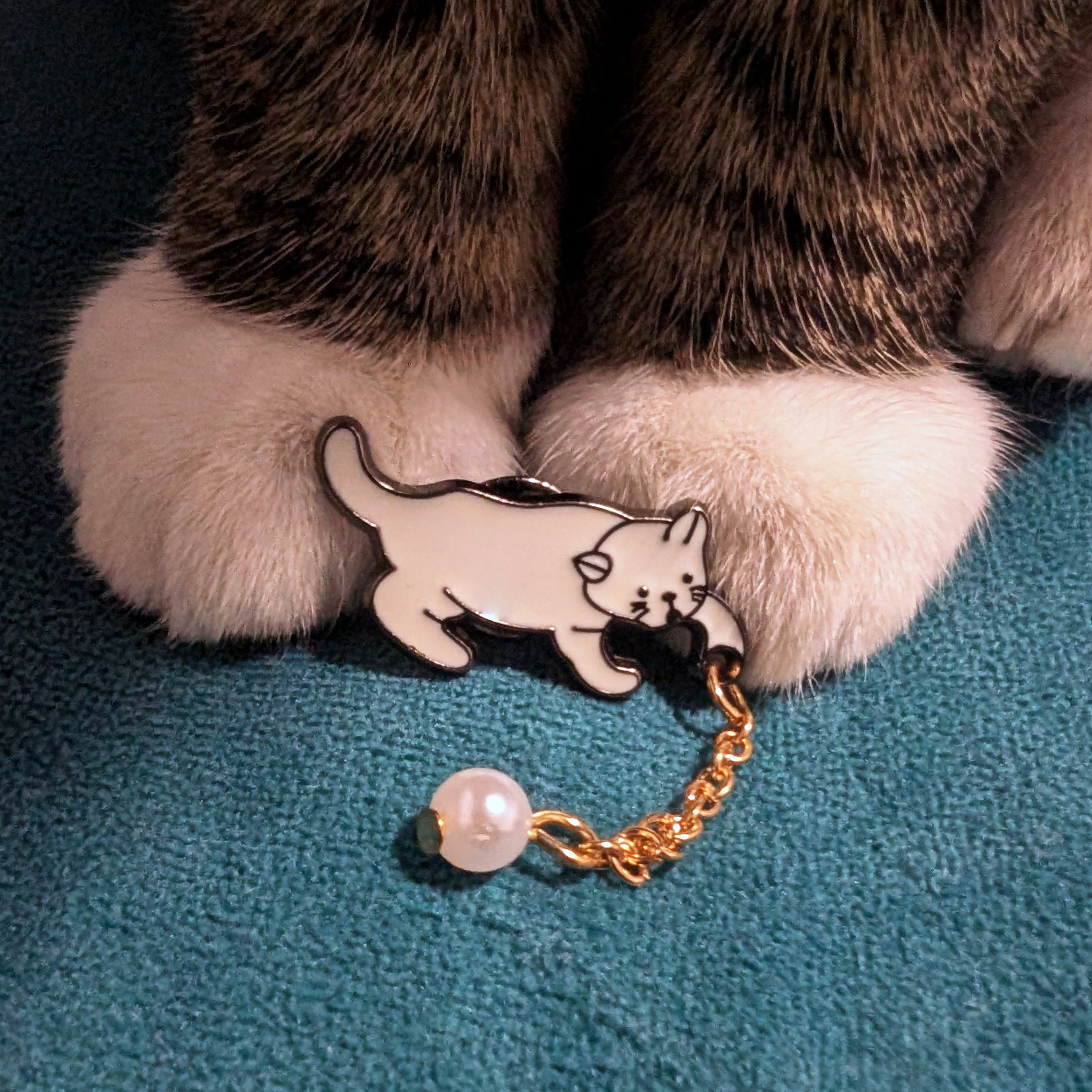 A cute white cat enamel pin with a gold chain and dangling pearl bead.