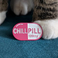 A pink and white capsule-shaped enamel pin with the text 'CHILLPILL 100mg' resting on a teal blue textured surface, with a cat's paws visible in the background.