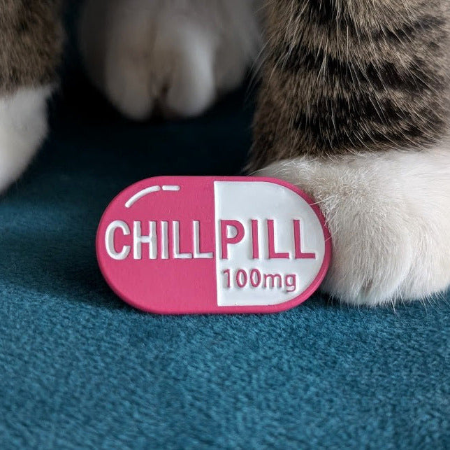 A pink and white capsule-shaped enamel pin with the text 'CHILLPILL 100mg' resting on a teal blue textured surface, with a cat's paws visible in the background.