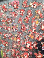 A dense collection of small red fox character charms or stickers arranged closely together on a dark surface. The characters have white and cream coloring with black outlines and various small accessories, photographed from above.