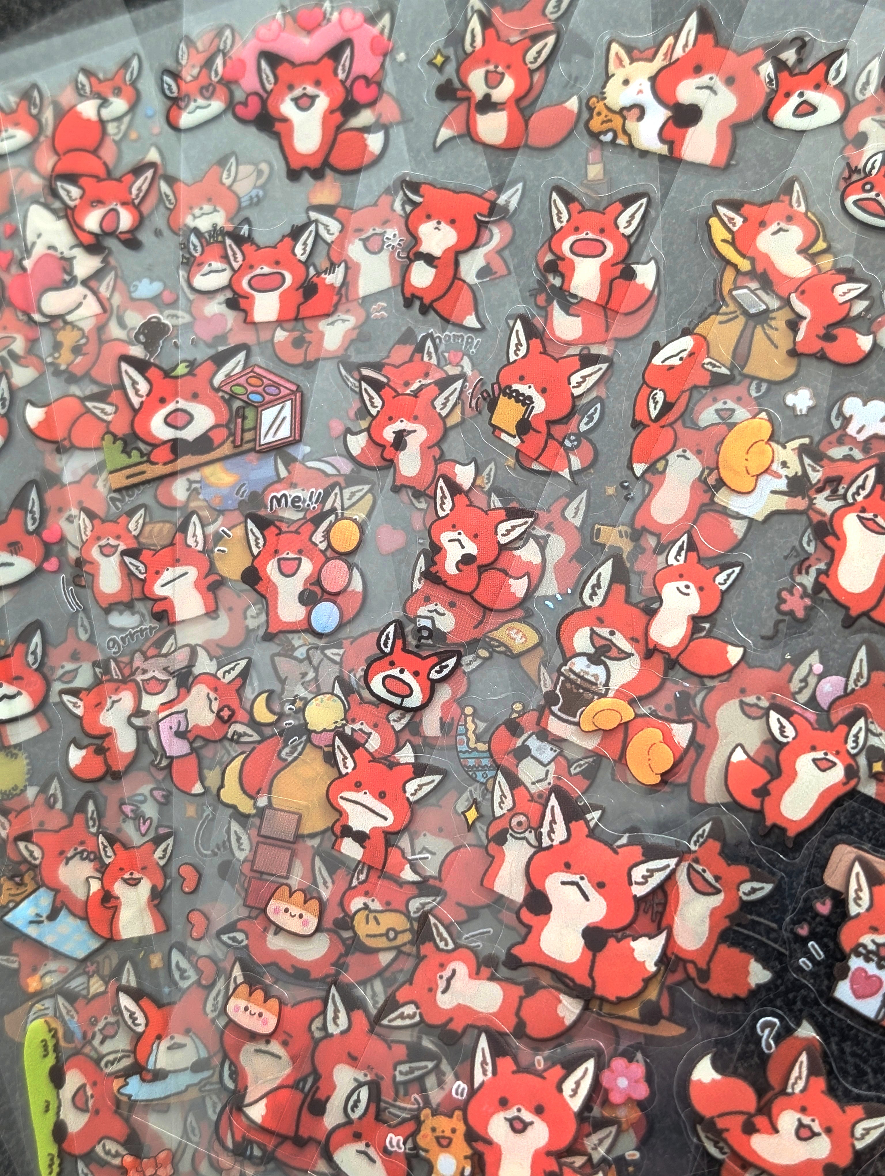 A dense collection of small red fox character charms or stickers arranged closely together on a dark surface. The characters have white and cream coloring with black outlines and various small accessories, photographed from above.