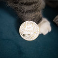 A round white enamel pin with gold-toned floral detailing and the engraved text 'I AM ENOUGH', shown next to a cat's paw on a dark teal fabric surface.