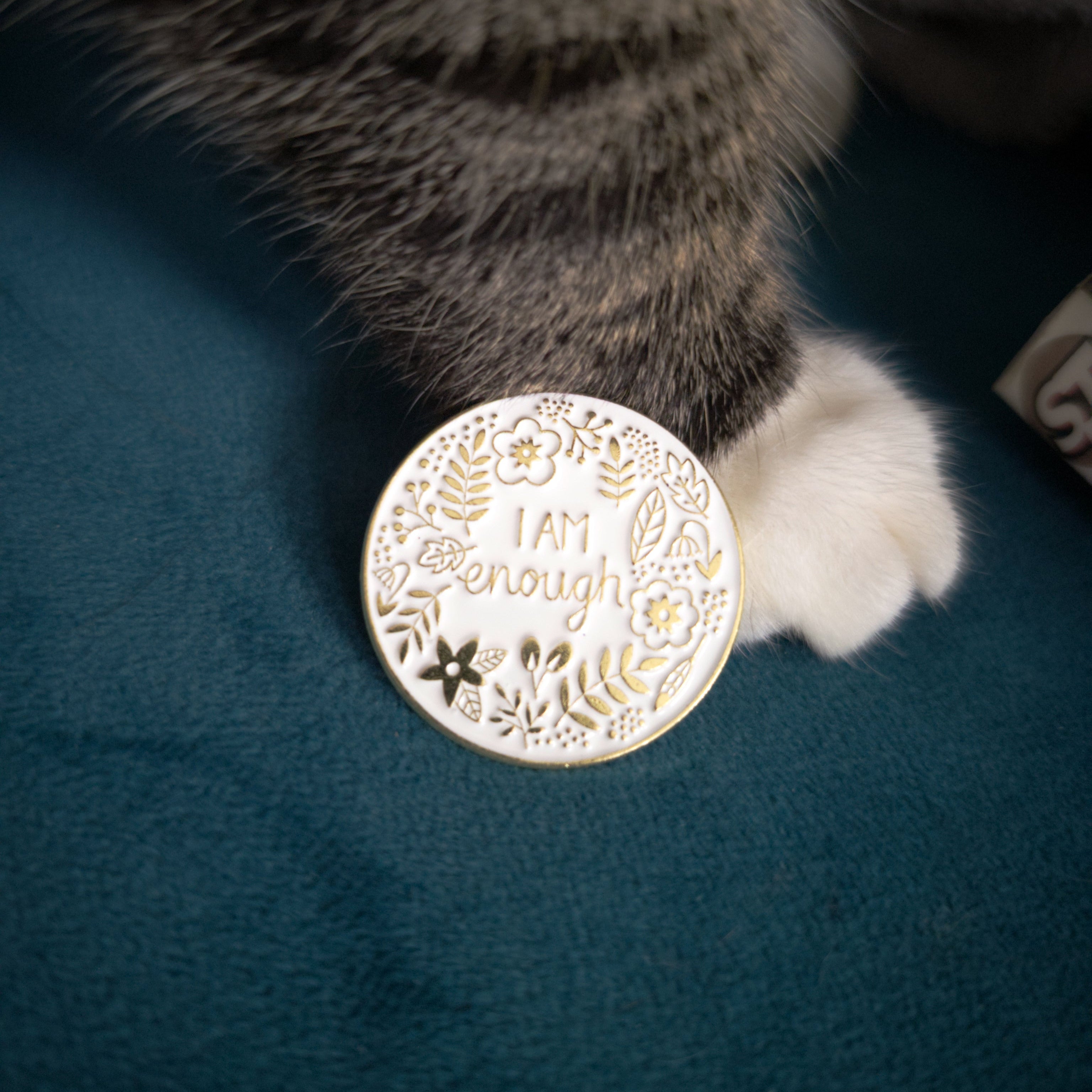 A round white enamel pin with gold-toned floral detailing and the engraved text 'I AM ENOUGH', shown next to a cat's paw on a dark teal fabric surface.