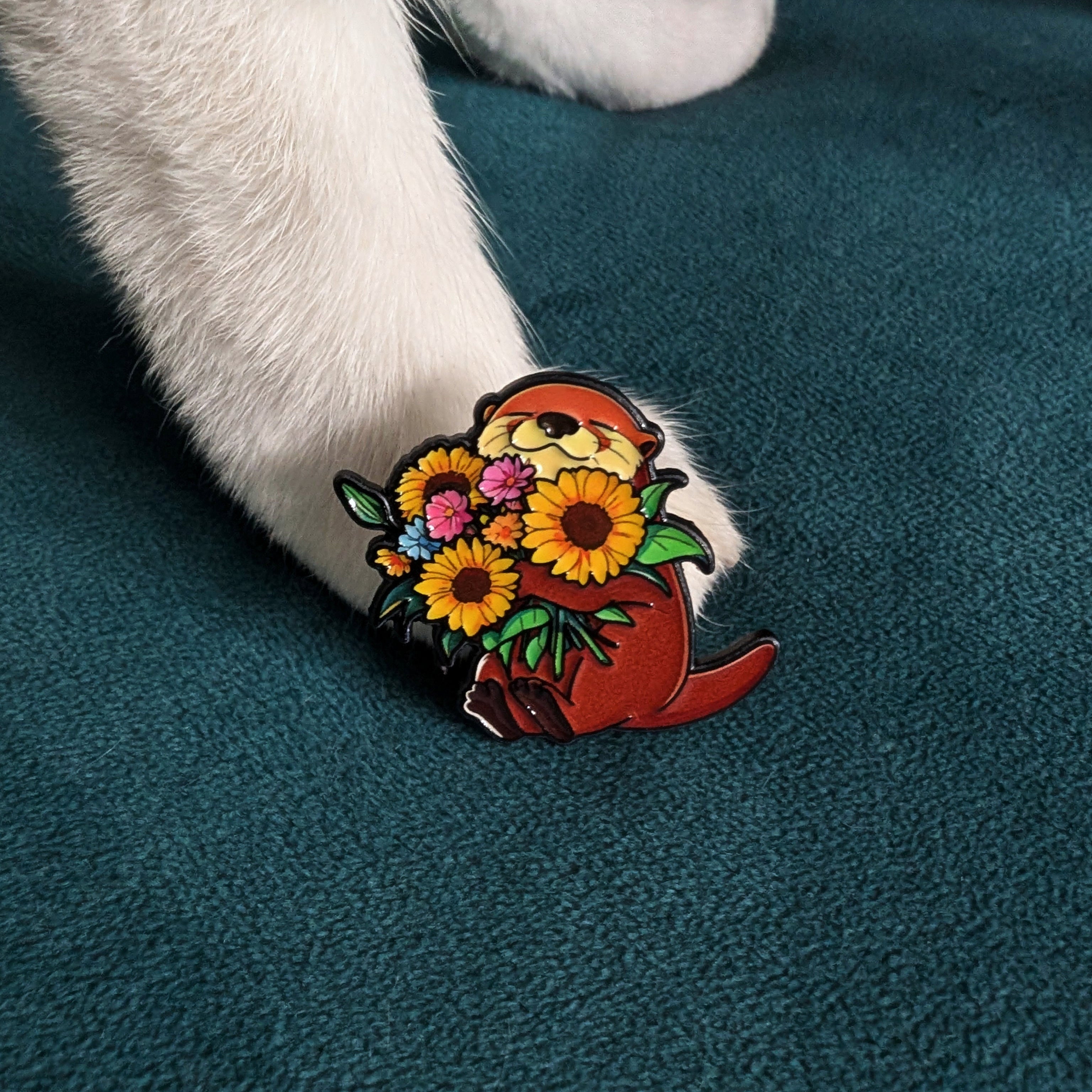 A small enamel pin shaped like an otter holding a bouquet of yellow sunflowers and pink flowers, shown clipped onto a white cat's paw resting on a dark teal textured blanket.