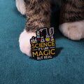 Enamel pin placed on the paw. The pin is black with white and yellow text reading 'SCIENCE IT'S LIKE MAGIC BUT REAL'.