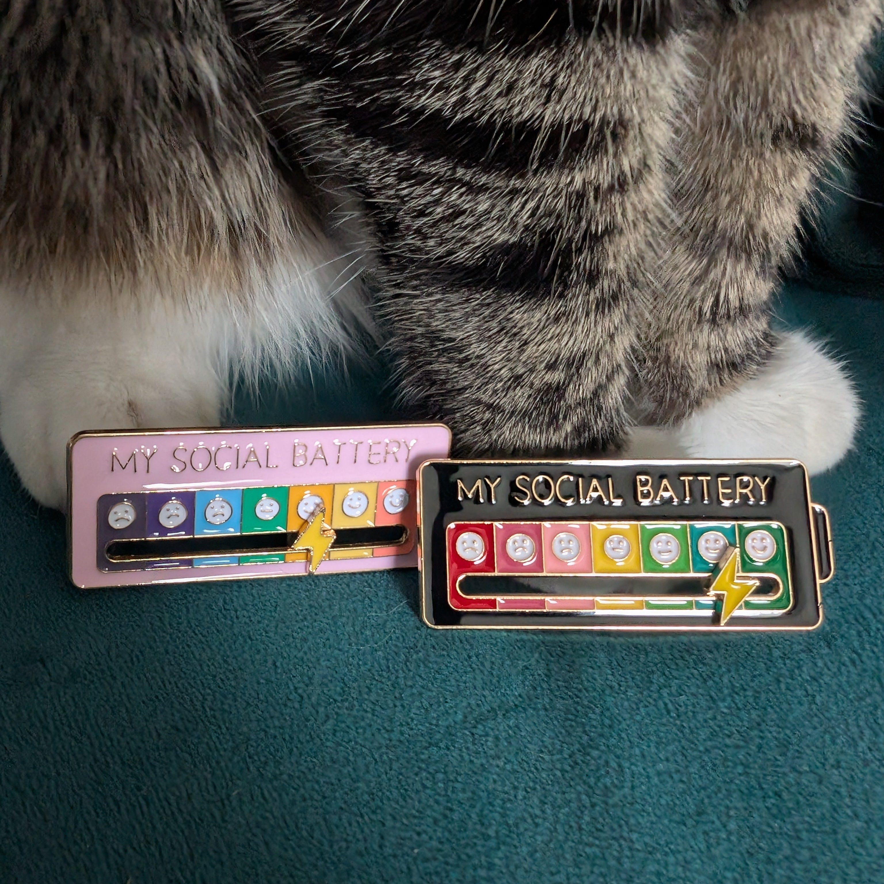 Two enamel pins shaped like battery chargers with the text 'MY SOCIAL BATTERY' are displayed on a dark teal surface, with a gray and white tabby cat's paws resting above them.
