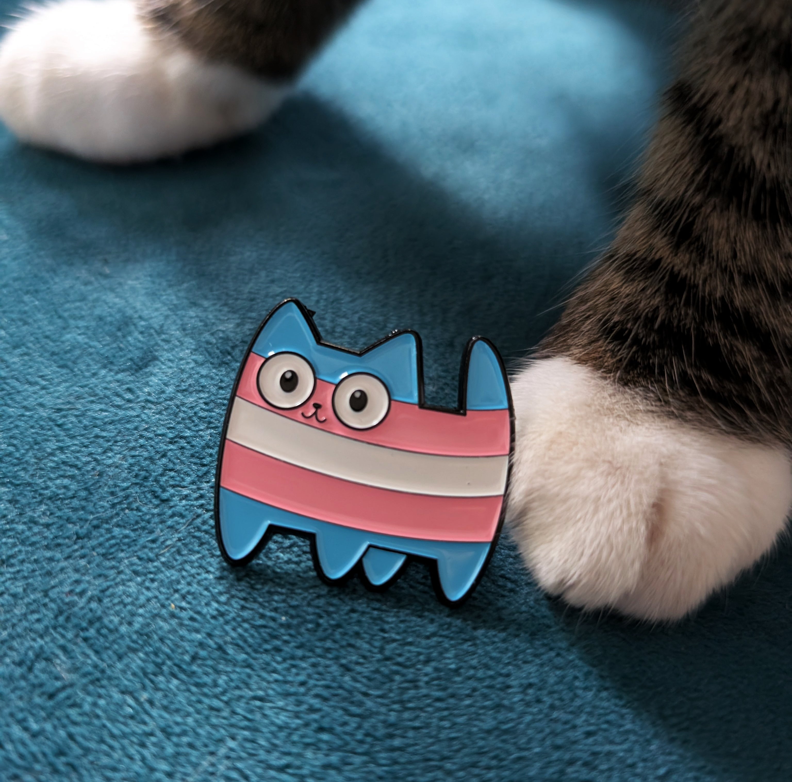 Cute cartoon cat figurine with a transgender pride flag design next to a cat's paw on a blue surface.