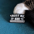 White enamel pin that reads 'TRUST ME, I'M A PROGRAMMER'.