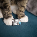 Cat's paws on a blue surface with a 'VERIFIED IDIOT' pin
