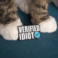 A close-up of a cat's paws standing on a dark teal blanket next to a small enamel pin that reads 'VERIFIED IDIOT' with a blue checkmark.