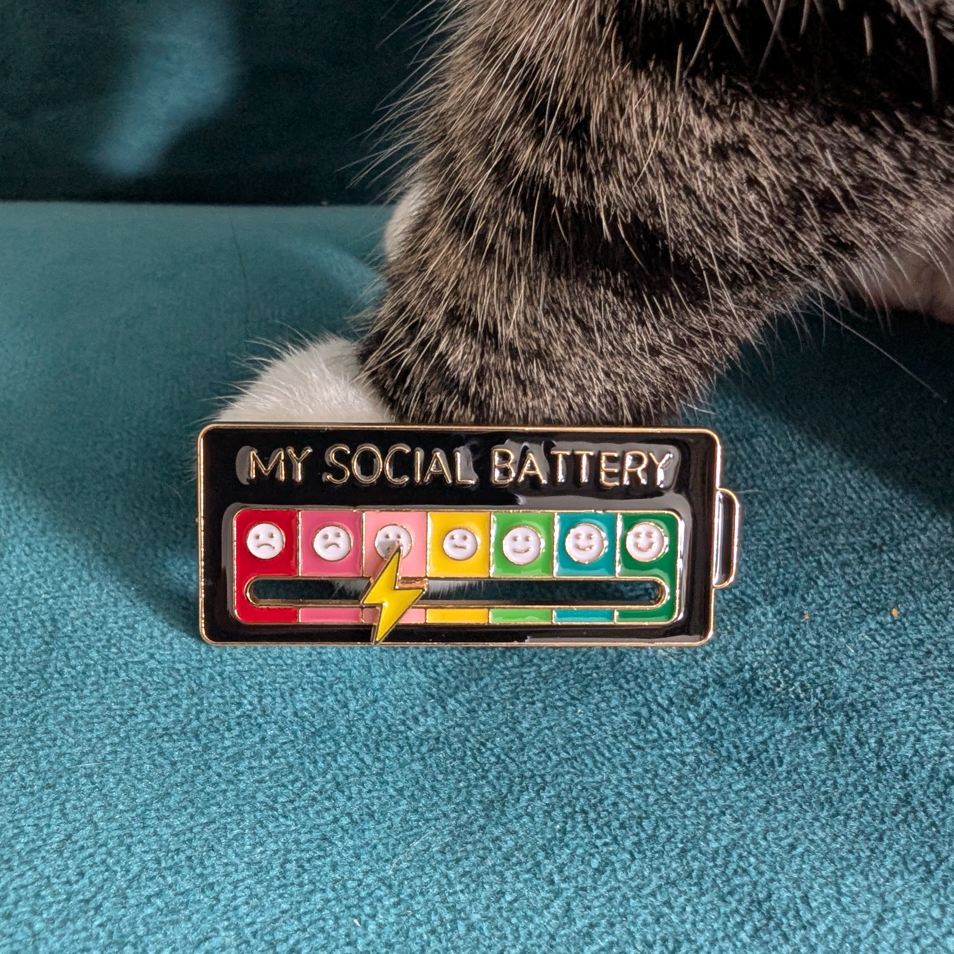 A rectangular enamel pin with the text 'MY SOCIAL BATTERY' above a battery-style graphic with red, pink, yellow, and green segments and cartoon faces, placed on a teal textured surface with a cat paw resting near it.