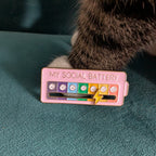 A pink enamel pin shaped like a battery labeled 'MY SOCIAL BATTERY' with colorful emoji-style indicators and a yellow lightning bolt, shown on a teal textured surface with part of a cat's paw and body visible.